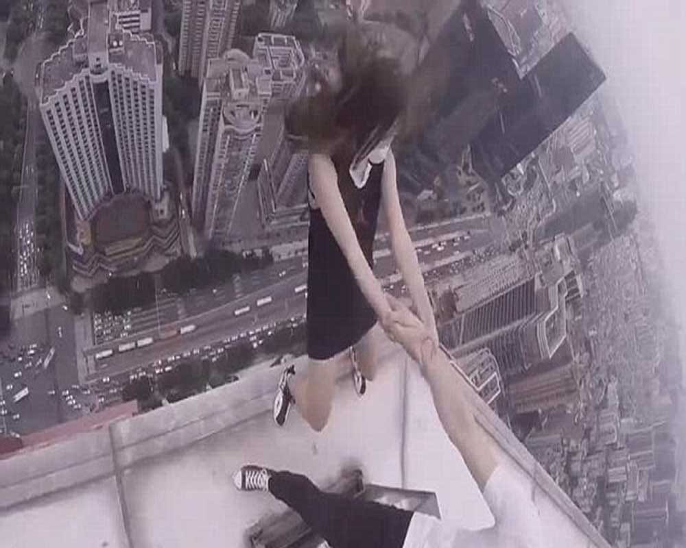 woman dangles from the edge of a skycraper as her boyfriend clings onto one arm 