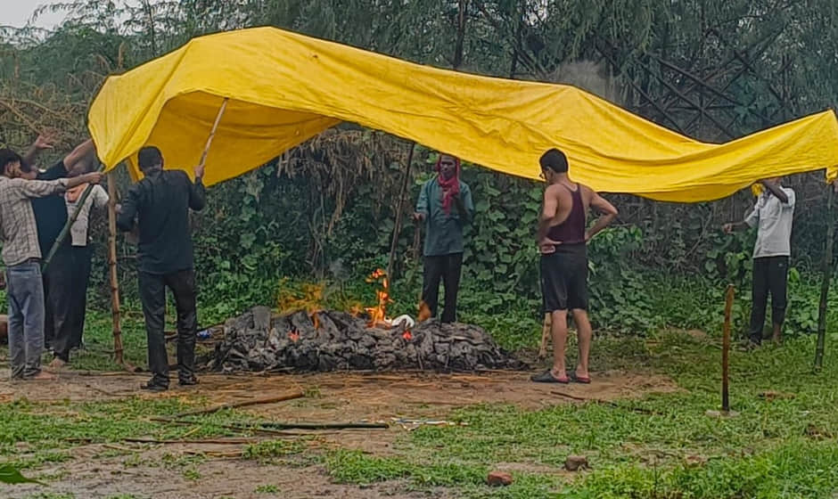 Funeral held under a makeshift tarpaulin amidst rain; two young children lose their father