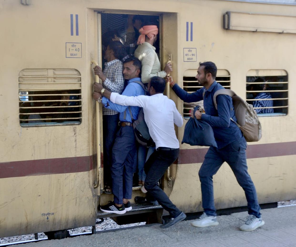 There is a huge crowd in trains... passengers are travelling by hanging on the doors