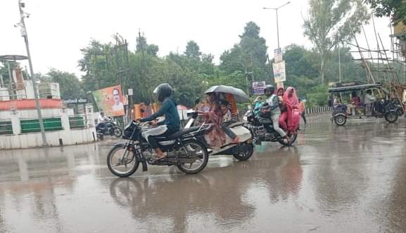 अचानक हुई बारिश के चलते नगर के टीडी कालेज चौराह स्कूटी पर छाता लेकर अपने गंतव्य को जाती महिला व