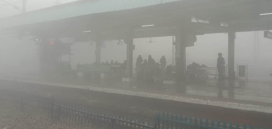 कोहरे के बीच प्लेटफार्म पर गाड़ियों के इंतजार में बैठे यात्रा।- संवाद।