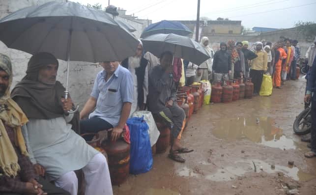 गांव जरोठी स्थित गैस गोदाम पर गैस सिलिंडर लेने के लिए बारिश में छाता लगाकर अपने सिलिंडरों पर की