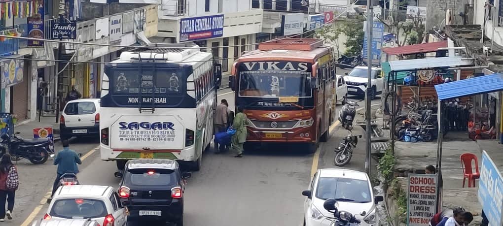 मैहरे बाजार में आमने सामने खड़ी बसों के बीच से गुजरते हुए लोग। संवाद