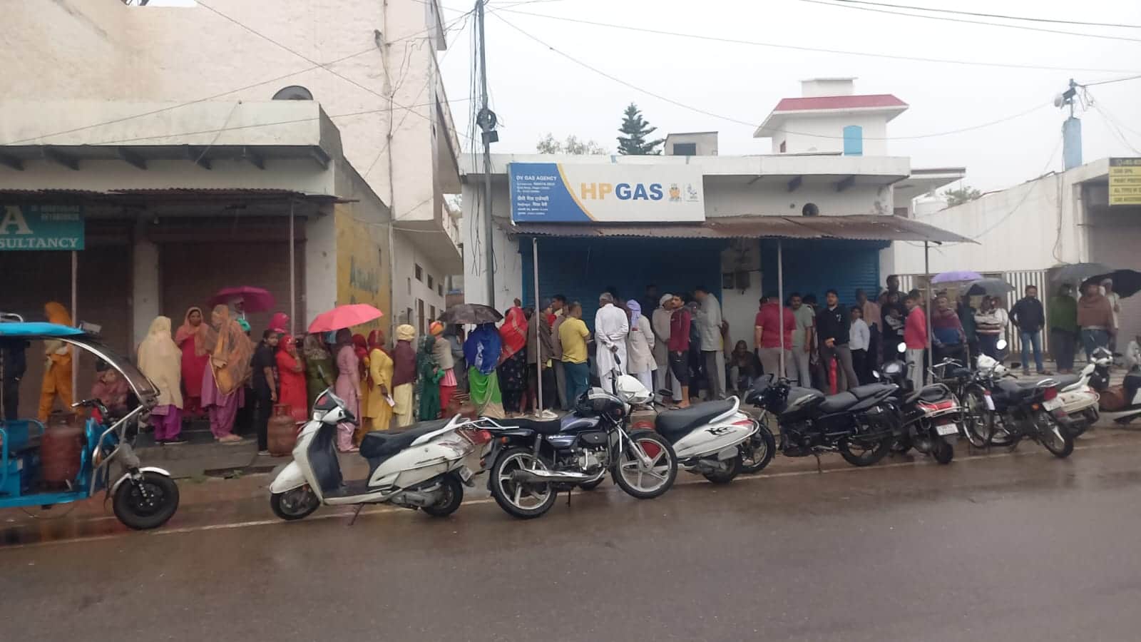 20jjrp14- कोसली रोड स्थित एचपी की गैस एजेंसी के बाहर बारिश में भी छाता लेकर सिलिंडर पर्ची के ल
