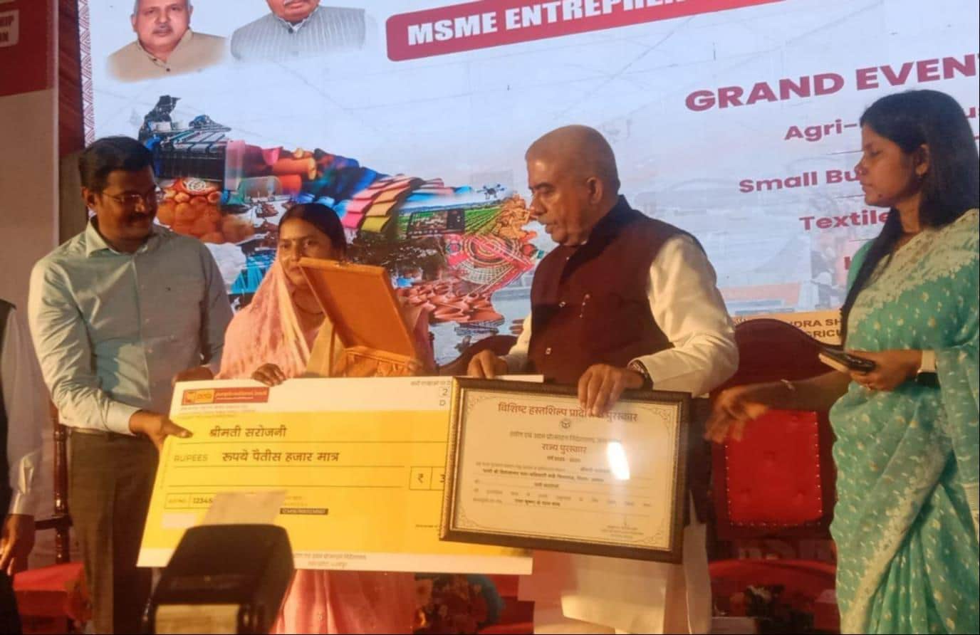 फोटो-1-कैबिनेट मंत्री राकेश सचान से प्रतीकात्मक चेक व प्रमाणपत्र लेतीं सरोजनी देवी। स्रोत: स्वय