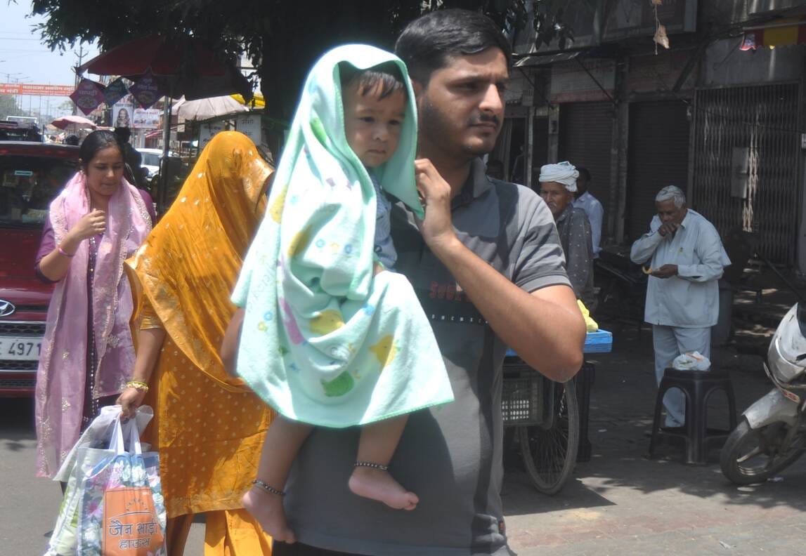 गढ रोड पर धूप में बच्चे को कपड़े से ढककर ले जाते युवक। संवाद