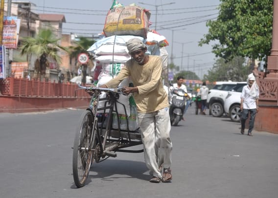 दोपहर के समय धूप में सामान से भरे रिक्शा को पैदल खींचकर ले जाते बुजुर्ग व्यक्ति। संवाद