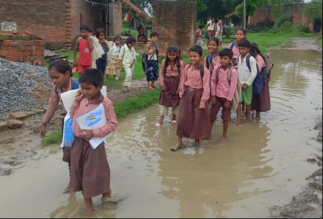 संदना में गुजरेहटा मार्ग पर भरे पानी के बीच से निकलते स्कूली बच्चे। संवाद