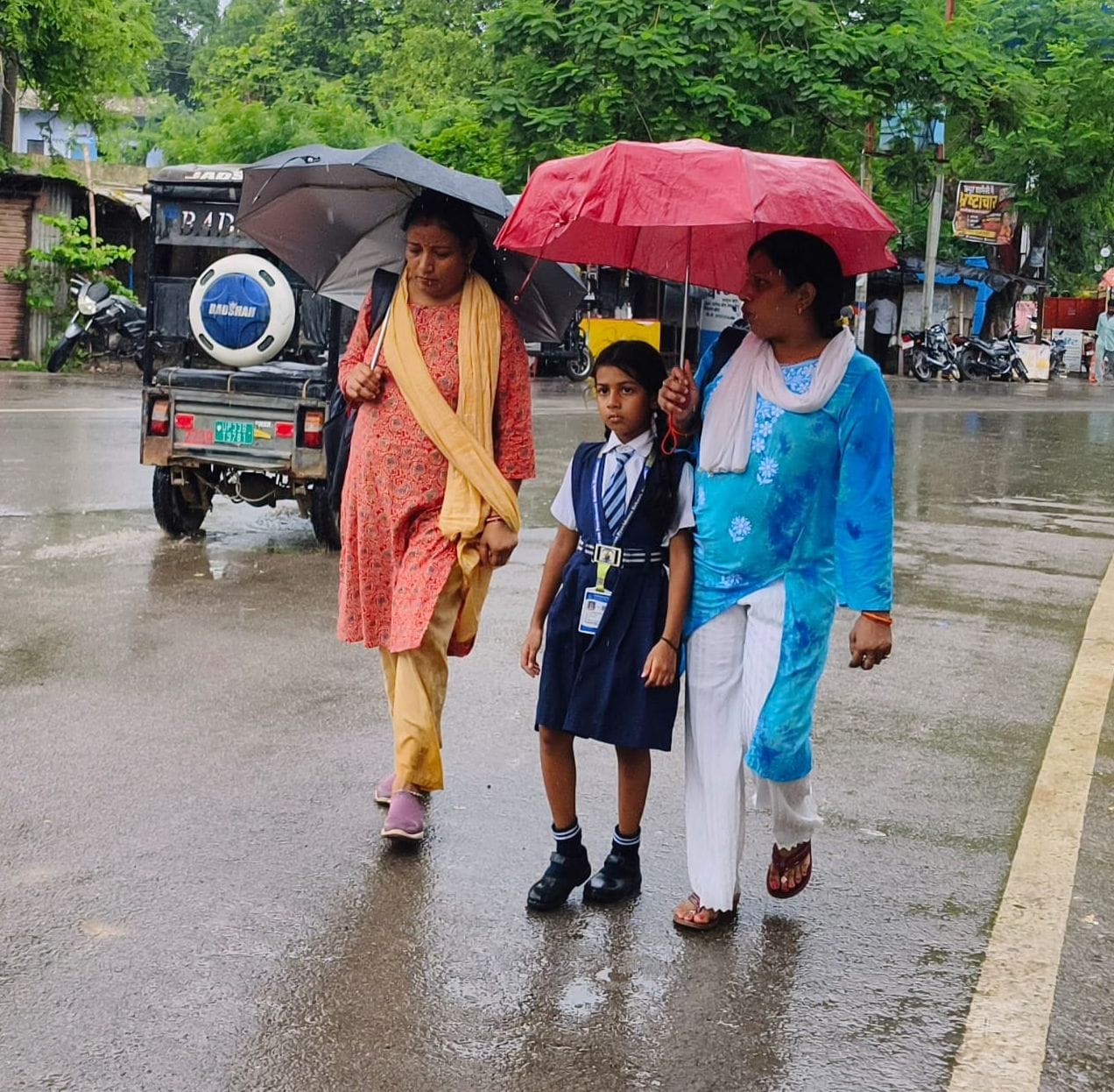 बारिश के बीच छाता लागाकर बच्ची के साथ निकलती महिलाएं। 