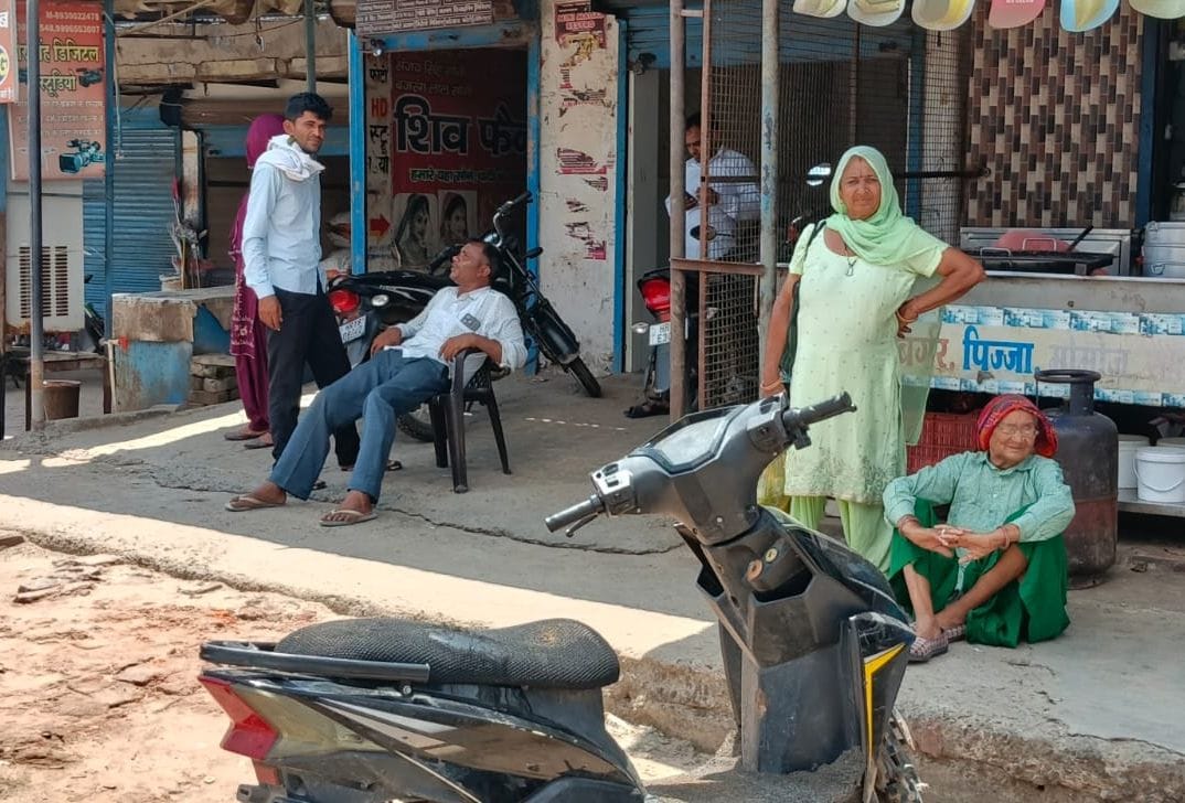 ढिगावामंडी में दूकानों के बाहर  खड़े होकर बस का इंतजार करते यात्री।