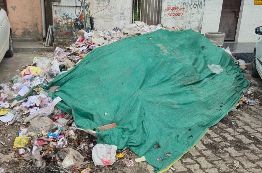 शहर में नेहरू पार्क के बाहर  सड़क के किनारे पड़ा कूड़ा कचरा पर ढ़का तिरपाल।