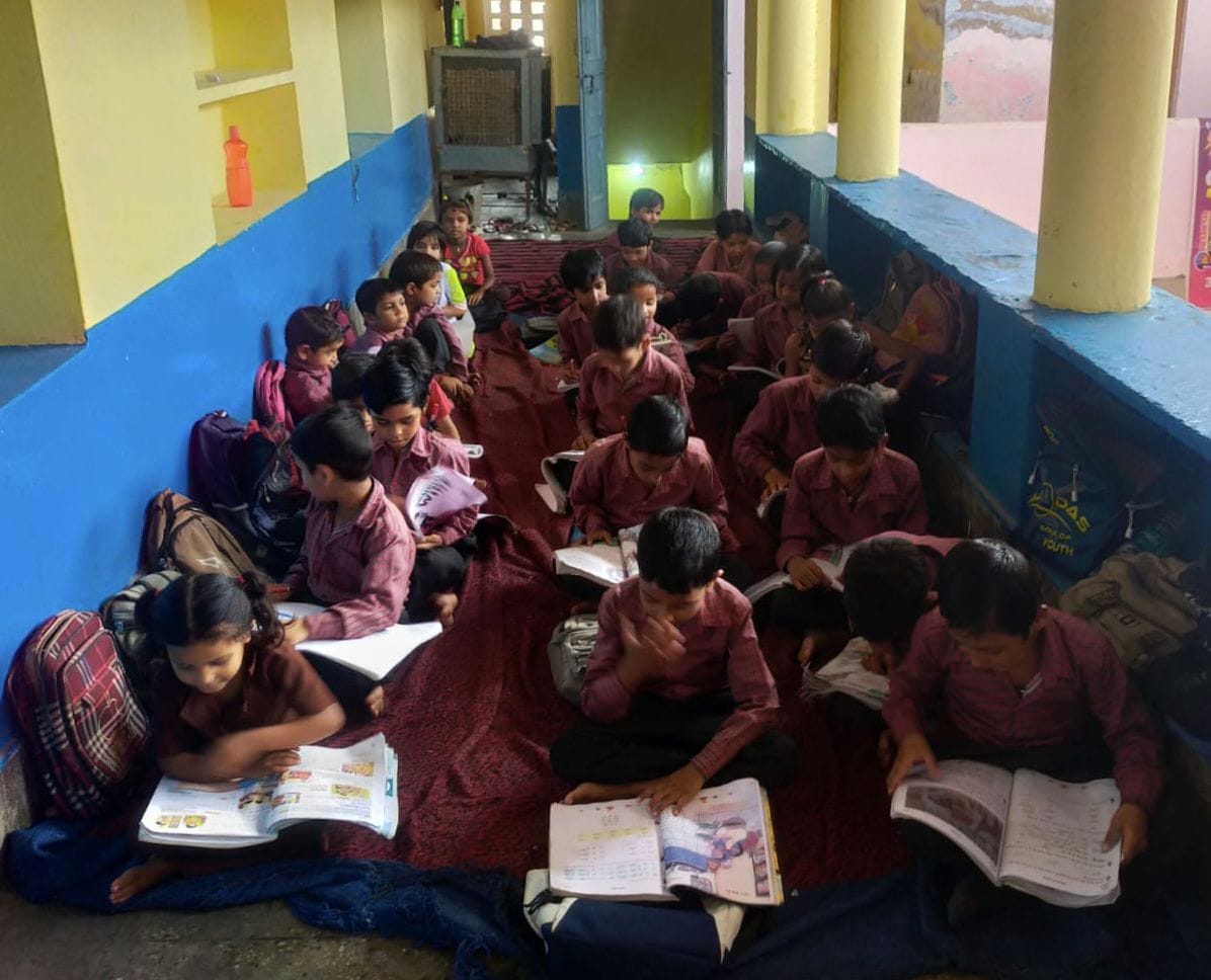 Dual desks were expected, yet young children are studying while sitting on jute mats.