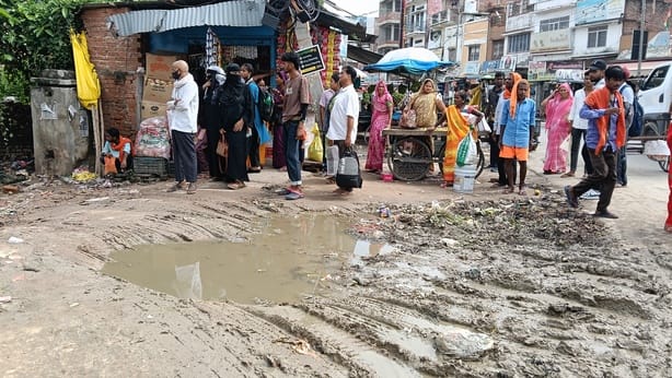 Passengers stand in the mud and sun waiting for the bus.