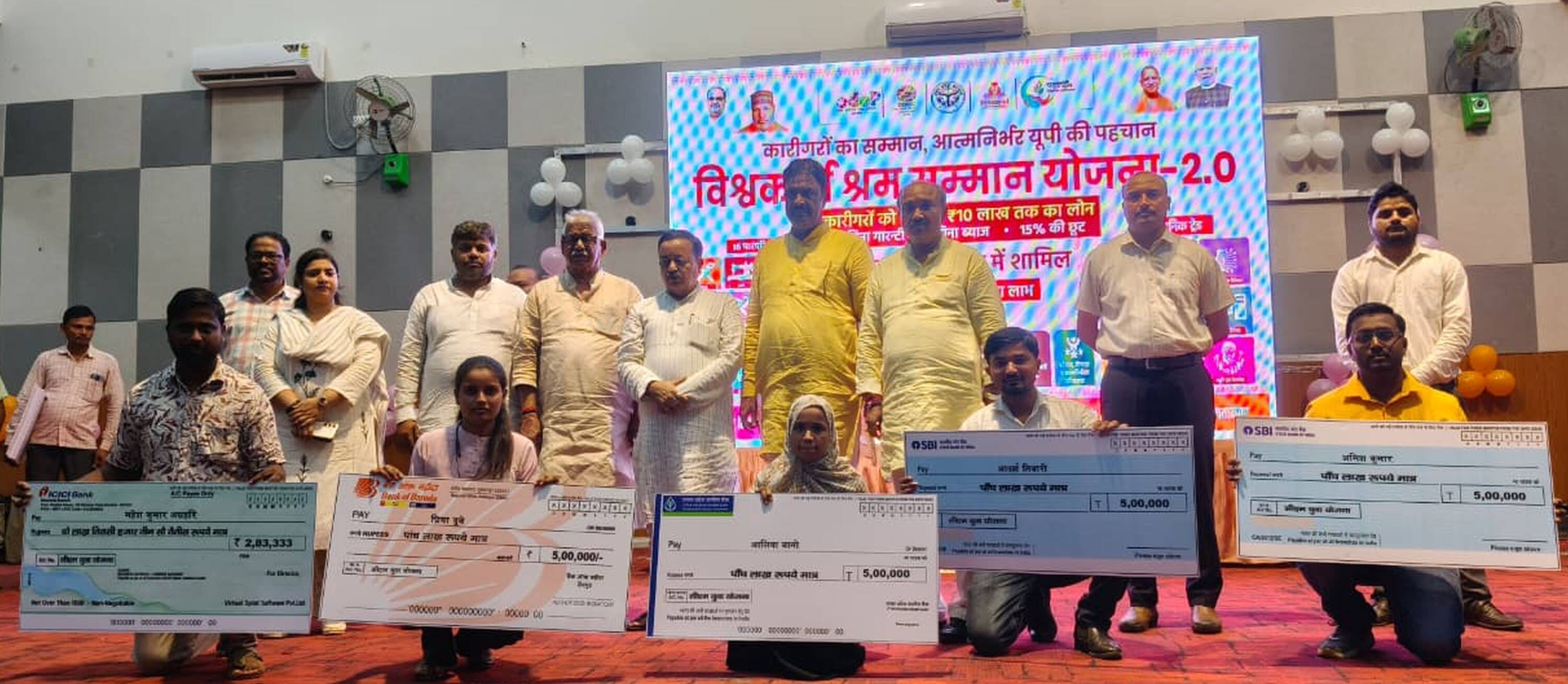 पं. राम नरेश त्रिपाठी सभागार में प्रतीकात्मक चेक दिखाते लाभार्थी। स्रोत- सूचना विभाग