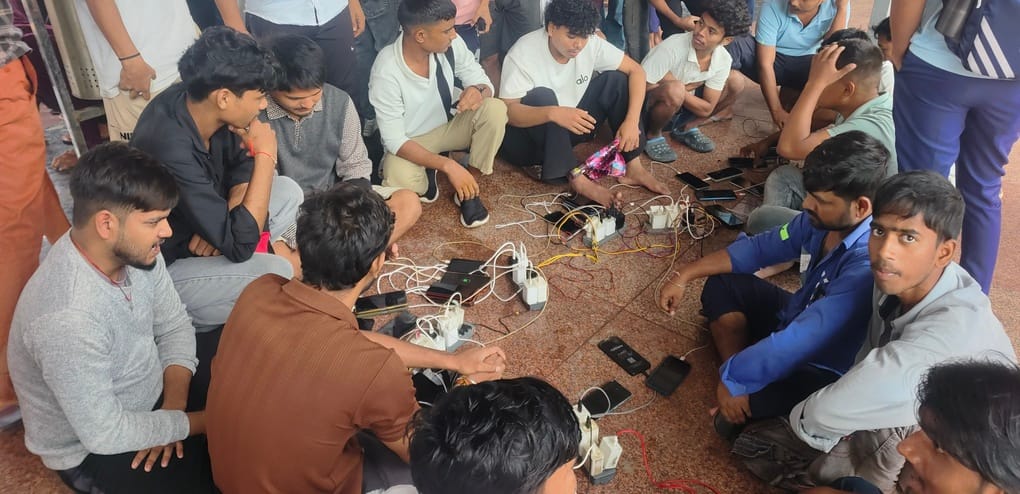 रेलवे स्टेशन पर जमीन पर एक्स्ट्रा बिजली का बोर्ड लगाकर बड़ी संख्या में एकत्र होकर मोबाइल चार्जि